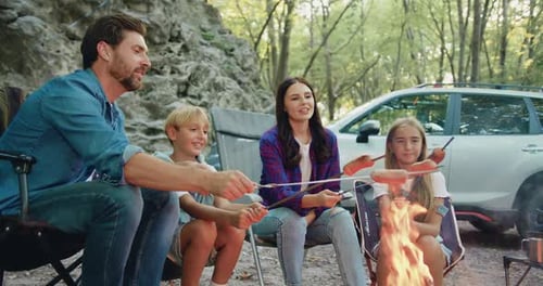 Smiling family father mother and son daughter roast sausage over campfire during picnic in forest