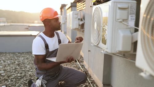 Man Inspecting Air Conditioning Units with Laptop on Rooftop