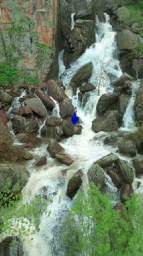 A Woman in a Flowing Blue Dress Poses Gracefully Near Cascading Waterfall Amid Rugged Rocks