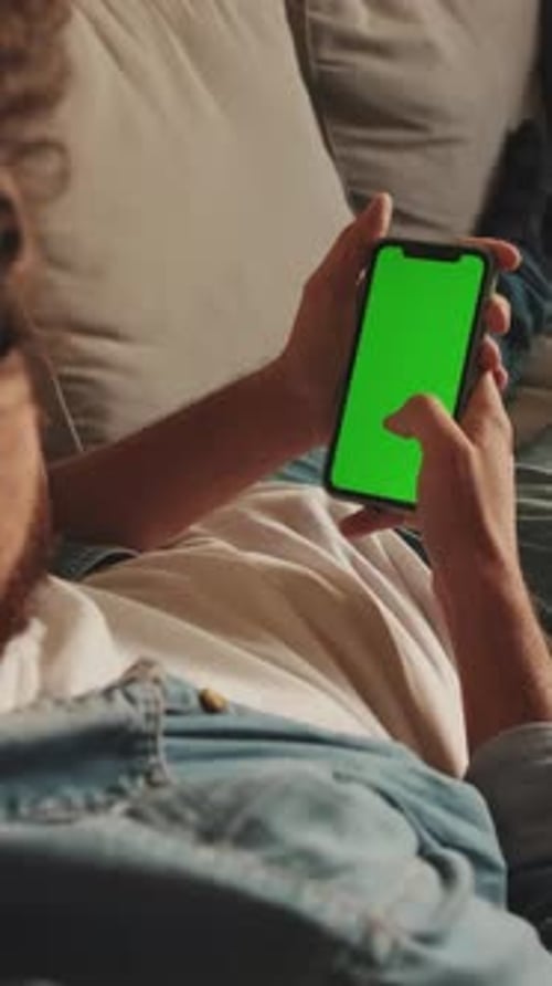 Young man lying on sofa uses mobile phone with green mock-up screen chroma key