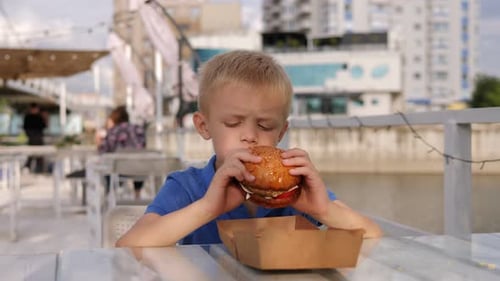 A Little Boy Eats a Big Burger in an Outdoor Restaurant in Summer