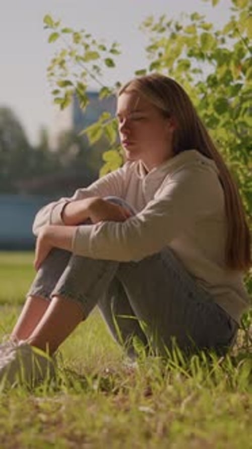 Thoughtful Woman Sitting on Grass in Sunlight with Urban Stadium in Background