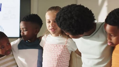 Diverse children linking arms, talking after display cue in class, girl in pink dress presenting