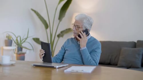Senior Man Talking on Phone While Trying to Turn on Tablet