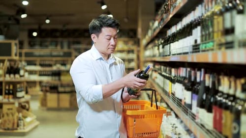 Asian man chooses wine in liquor store or supermarket. The male buyer examines the bottle of wine