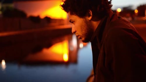 Serious Looking Young Man Drinks Can While Standing Over Water at Night in Orange Lighting