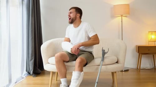 Injured Man with Casts Sits on Couch in Pain
