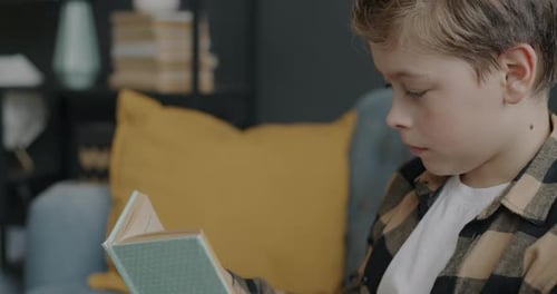 Porträt eines klugen Kindes, das ein interessantes Buch liest, das auf einer bequemen Couch in der Wohnung sitzt