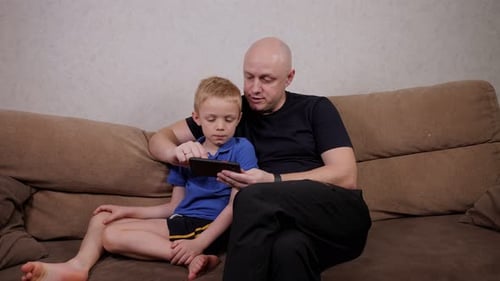 Happy Father and Son Play Together on a Tablet While Sitting at Home on the Sofa