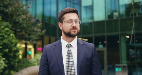 Portrait of serious and successful Caucasian businessman in suit and glasses, standing on the street