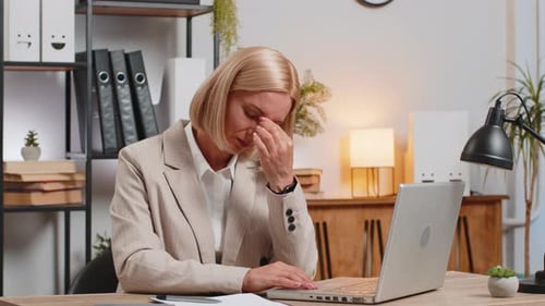 Exhausted Mature Businesswoman Girl Suffering From Headache While Using Laptop at Home Office Desk