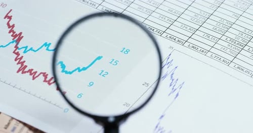 A hand with a magnifying glass passes over a financial newspaper and financial bag with a business