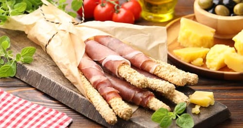 Tasty grissini breadsticks with prosciutto and cheese on wooden table, closeup. Camera moving right