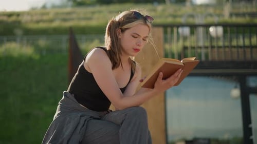 Woman Reading a Book Outdoors with Wind Blowing Through Her Hair on a Green Hill