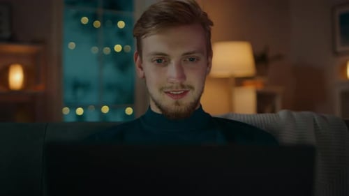 In the Evening Handsome Young Man at Home Sitting on a Couch in the Living Room Works on a Laptop C