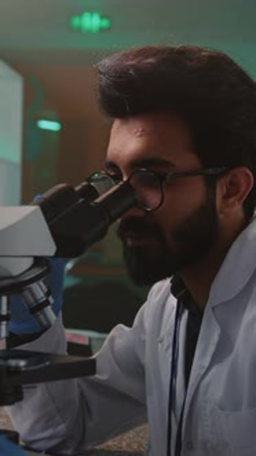 Retrato de un joven barbudo con gafas trabajando en el microscopio durante el examen de muestras en