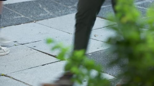 Auf dem gefliesten Bürgersteig sind vorbeifahrende Füße mit weißen Turnschuhen zu sehen, Fußgänger, die zur Arbeit gehen