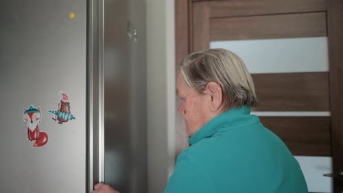 Elderly Woman in Green Sweatshirt Reaching to Open Refrigerator Door in Modern Kitchen