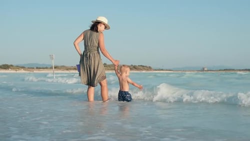 Cheerful Woman With Child Going To Water At Sea Beach. Happy Mother And Son Playing At Water Wave...