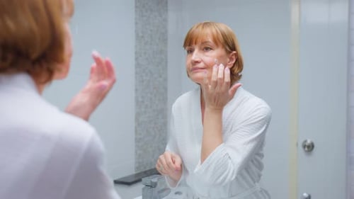 Woman Applies Cream to Face in Bathroom Mirror