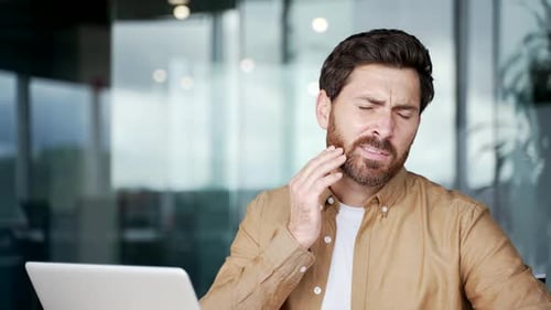 Upset businessman suffering from toothache while sitting at workplace at desk in business office.