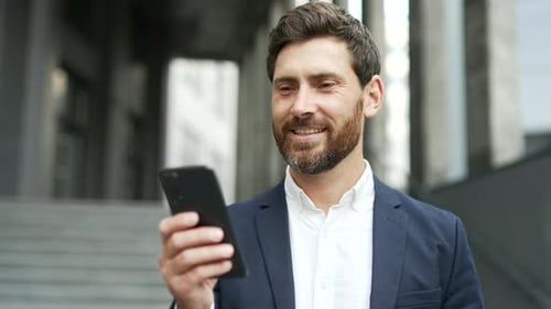 Smiling businessman in formal suit is using a mobile phone while standing on street near an office