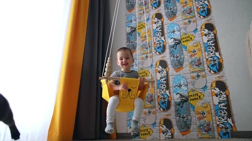 Smiling happy baby boy in a yellow swing. Adorable child waving his hand to the cat. Low angle view.
