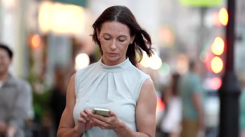 A Middleaged Woman with Short Brown Hair Stands on the Street with a Smartphone in Her Hands Her