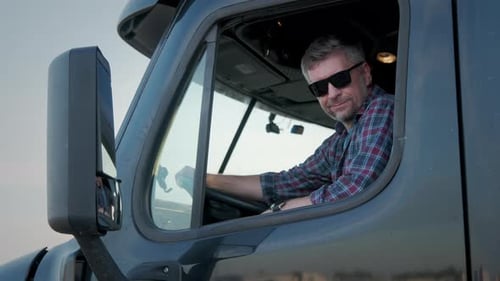 Professional Truck Driver Sits in His Truck and Behind Him Parked Long Haul SemiTruck Close Up
