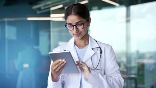 Close-up of focused young female doctor in white coat and glasses using digital tablet in modern