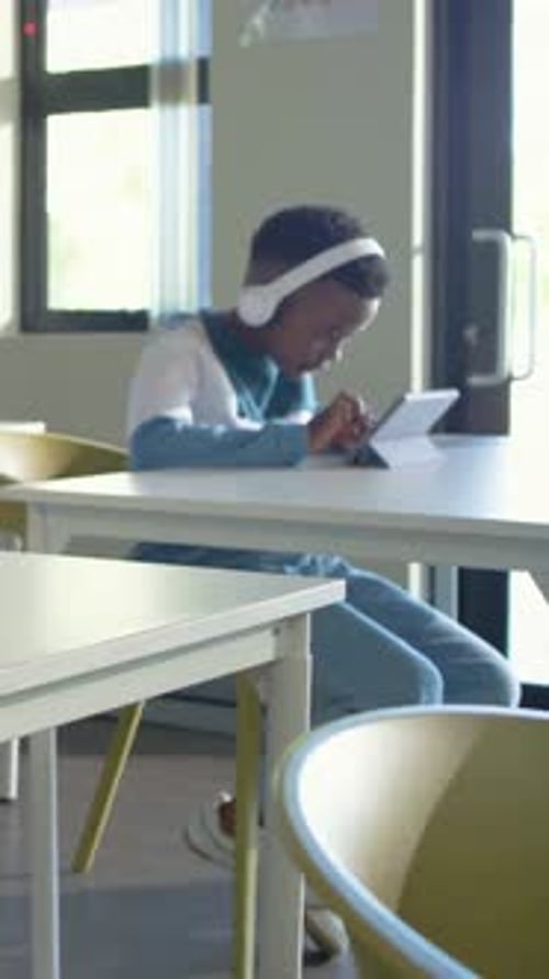 Vertical video: Tapping tablet, student with white headphones and blue top studying in classroom