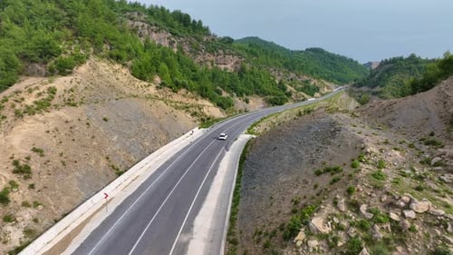 Aerial View of a White Car Driving on a Winding Highway Through Rocky Mountainous Terrain with Green