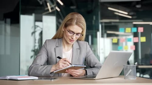 Woman Works at Laptop and Writes in Notebook