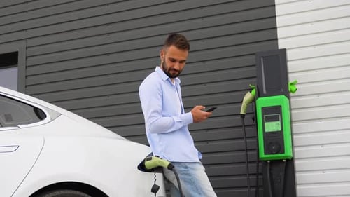 Stylish Man with Electric Car at Daytime Charging the Vehicle and Holding Smartphone