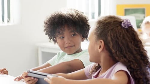 African American children sitting at school desk, girl sharing tablet, boy leaning and smiling