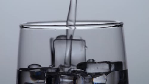 Close Up Of Drinking Water Being Poured Into A Glass With Ice Cubes On The Light White Background