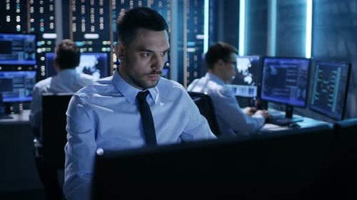Primer plano del ingeniero de operaciones trabajando en su computadora con múltiples pantallas en la sala de monitoreo.