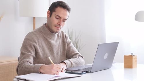 Man Writing in Notebook with Laptop at Desk