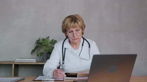 Adult Woman Doctor 60 Years Sitting at Desk in Office with a Laptop