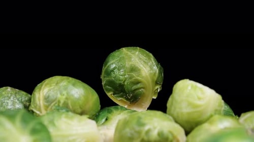 Scattered raw Brussels sprouts on a table against a black background, with one rotating in the dista