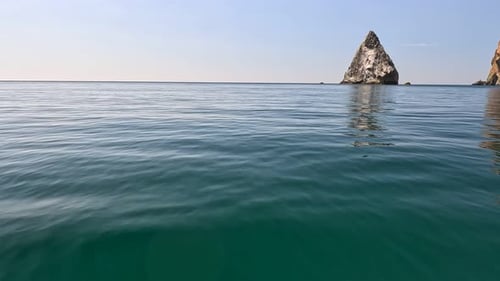 Sea Water Surface Camera Flies Over the Calm Azure Sea with Volcanic Rocky Shores on Background