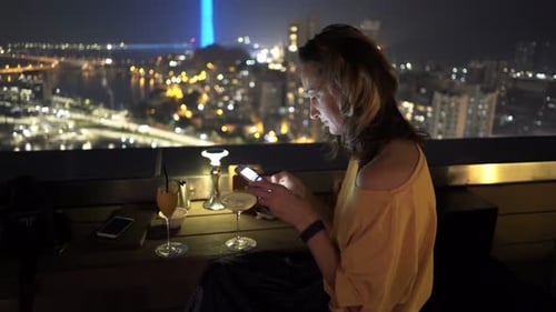 Young Woman with Smartphone Drinking Cocktail Sitting on Terrace in Bar At
