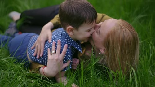 Slow Motion Loving Mother Tenderly Hugs and Kisses Her Little Son While While Lying on Green Grass