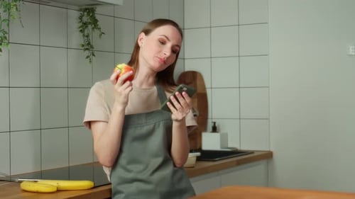 Woman Eating Apple and Using Smartphone in Kitchen