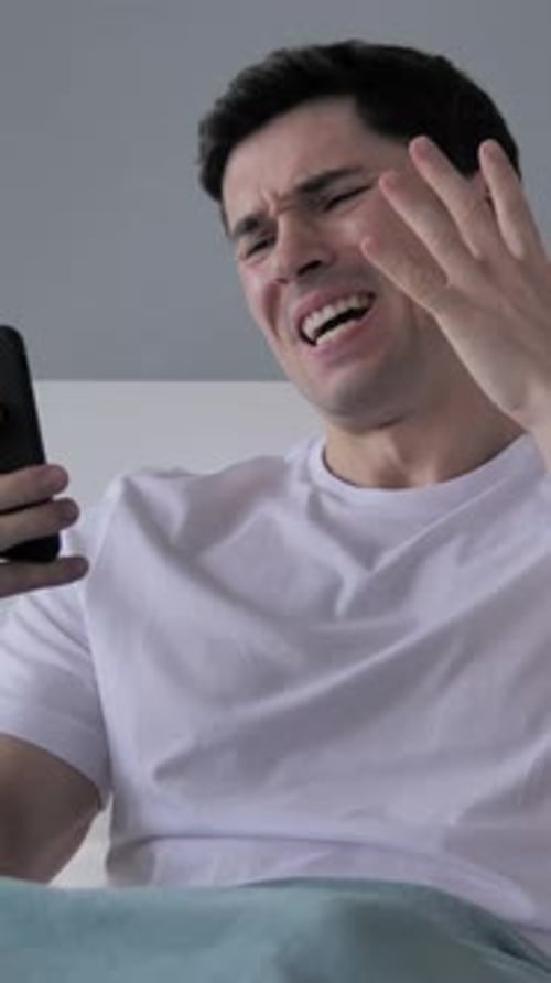 Young Man Reacting to Loss, Using Smartphone in Bed, Vertical Video
