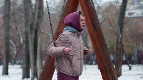 Young Woman Swinging in Snowy Park Holding Chains With Hoodie in Winter