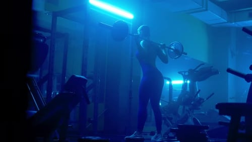 Rear View of a Dark Silhouette of an Athletic Fitness Woman is Squatting with Barbell in Dark Gym