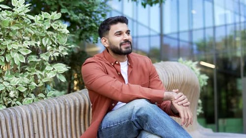 Happy carefree businessman in casual clothes sits on a bench on street near modern office building.