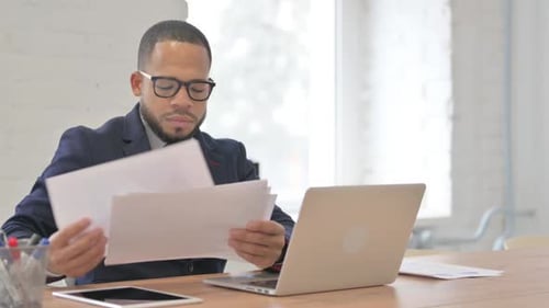 Man Reads Documents at Office Desk with Laptop