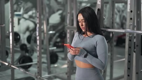 Woman Leaning Against Gym Rack Using Smartphone